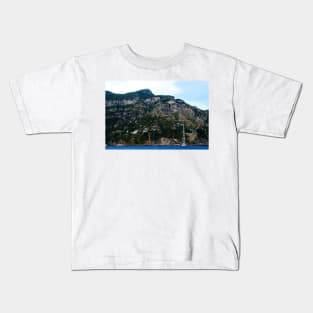 View in Amalfi coast at a huge mountain with rocky parts, greenery and buildings with a white boat underneath Kids T-Shirt